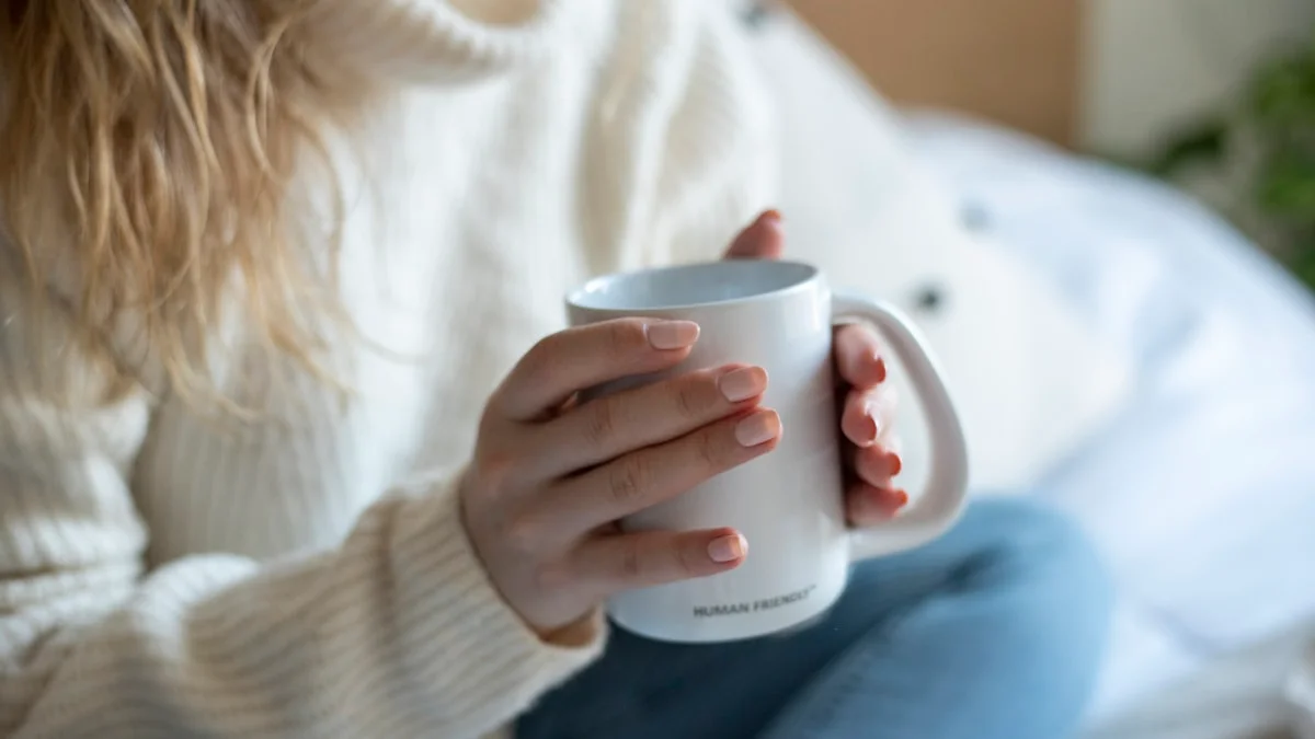 A woman in a cream knit sweater cradles a white mug in both hands in soft morning light - a Quiet Core Reset guide to 5-Minute Nervous System Regulation Practices for Real Life.