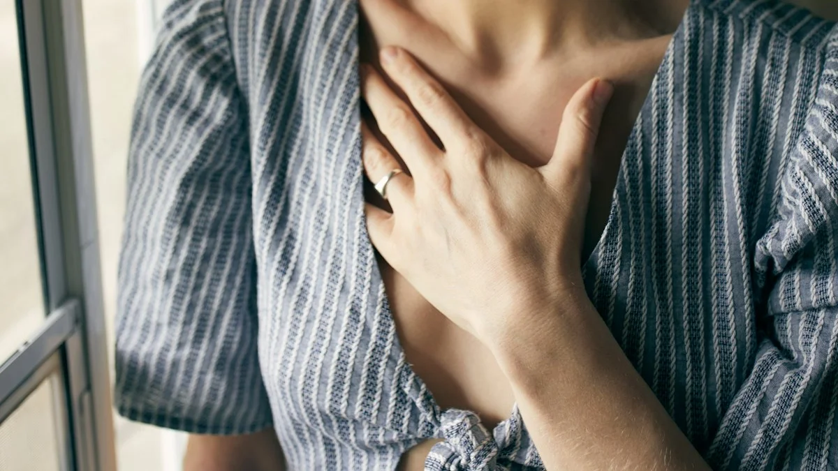 A woman by a bright window resting a hand flat against her chest, pausing to notice her body's signals - a Quiet Core Reset guide to 9 Signs of a Dysregulated Nervous System.