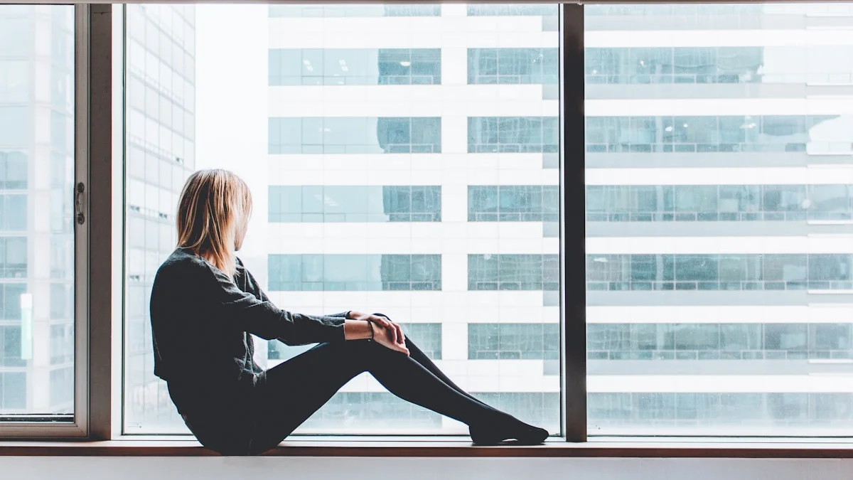 A woman taking a quiet pause on an office windowsill, hands resting on her knee, gazing out at the towers - a Quiet Core Reset guide to 90-Second Energy Check-In.