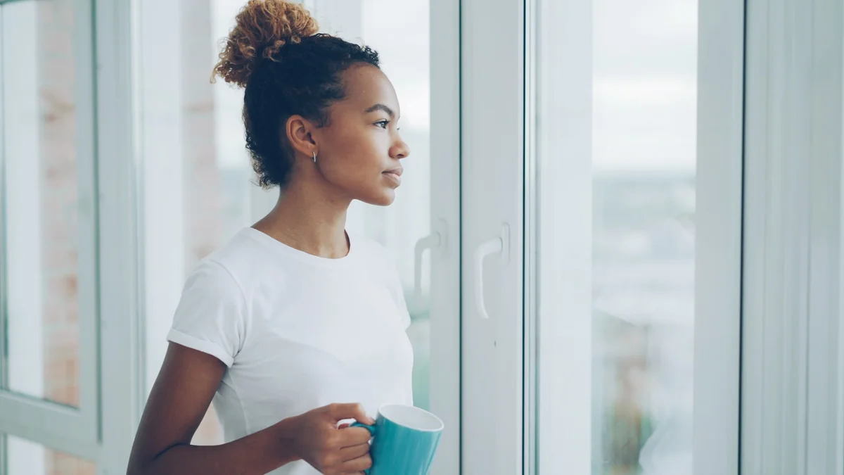 A woman holding a mug and looking out the window in quiet thought - a Quiet Core Reset guide to After-Work Decompression Window.