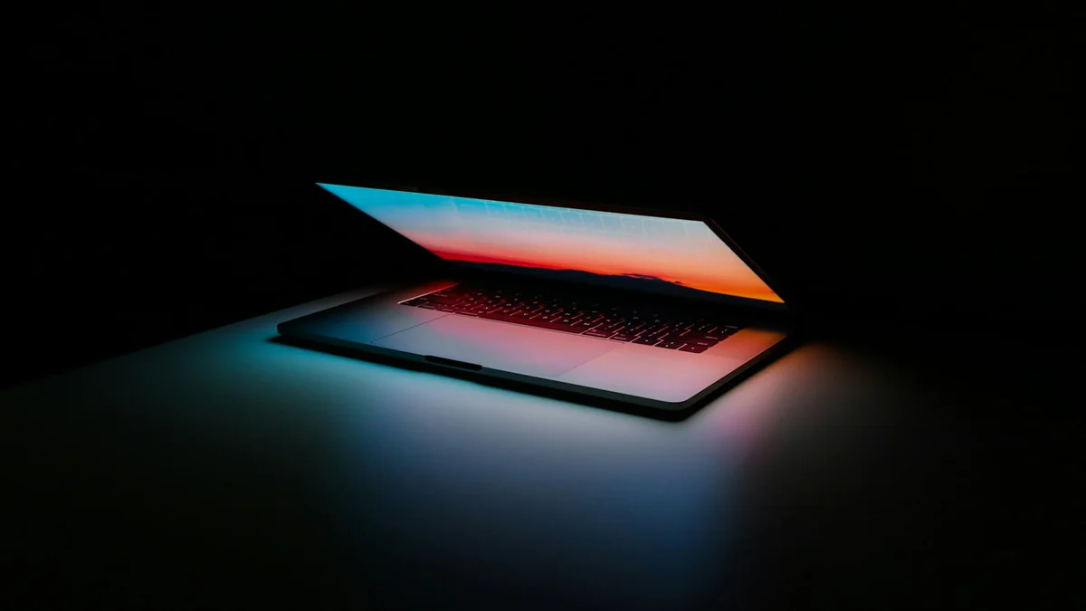 A laptop open in a dark room, its glowing screen reflecting colorful light onto the desk - a Quiet Core Reset guide to Attention Audit.
