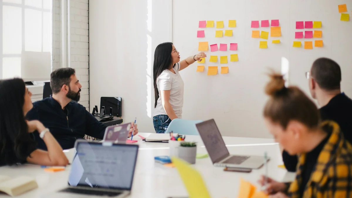 A woman placing sticky notes on a whiteboard while a team watches at a table, a planning session - a Quiet Core Reset guide to Boundaries for High Performers.