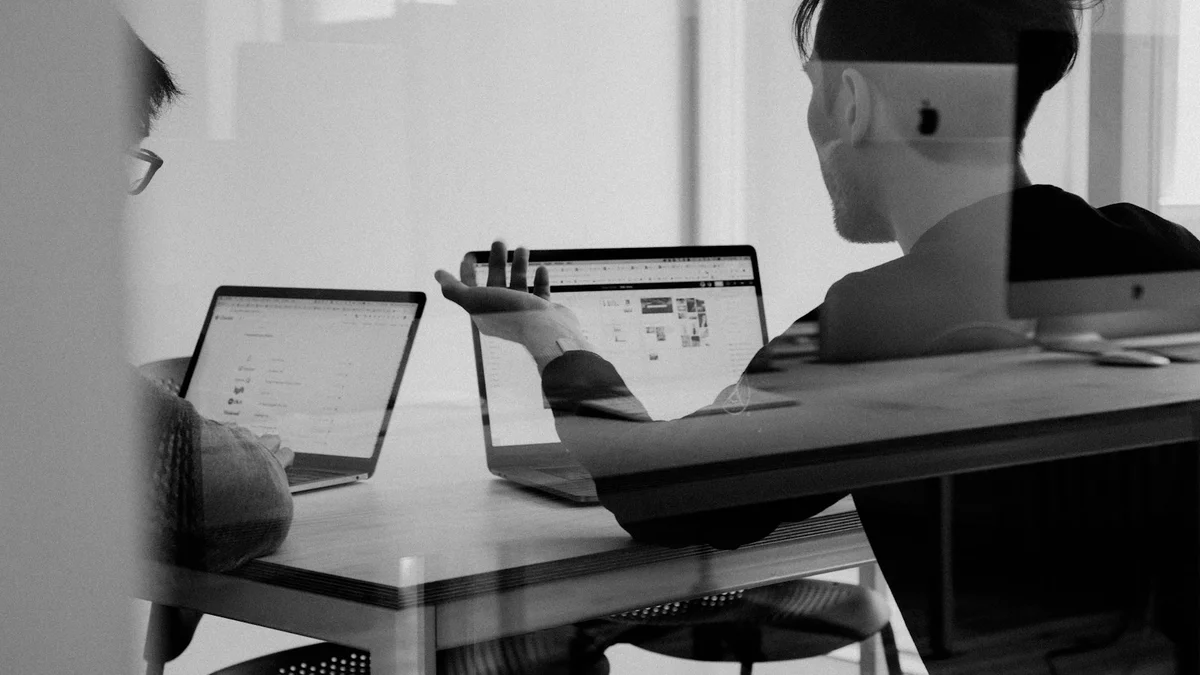 Two people in black-and-white discussing work at a desk with laptops, seen through glass - a Quiet Core Reset guide to Burnout Recovery.