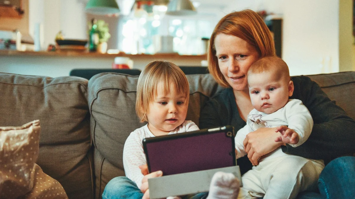 A mother sitting on a sofa holding a baby while a child looks at a tablet, a family at home - a Quiet Core Reset guide to Burnout Recovery for Parents.