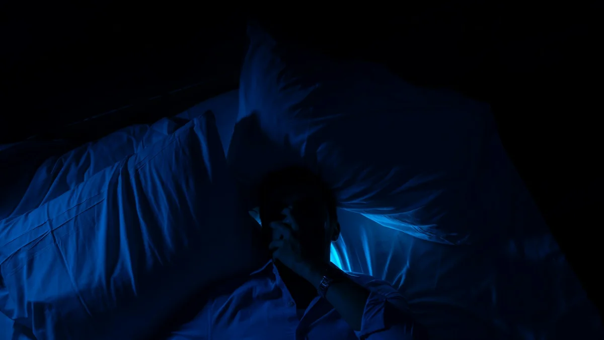 A person lying fully dressed on a bed in dark blue night light, one hand over their face - a Quiet Core Reset guide to Can't Rest Even When You're Burned Out? Here's Why.