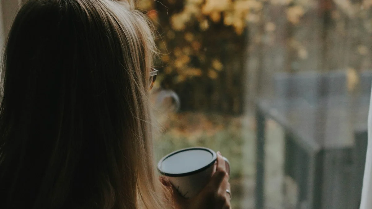 A woman holding a warm mug, gazing out a window at golden autumn trees - a Quiet Core Reset guide to Why Use Comparison as Motivation Backfires.