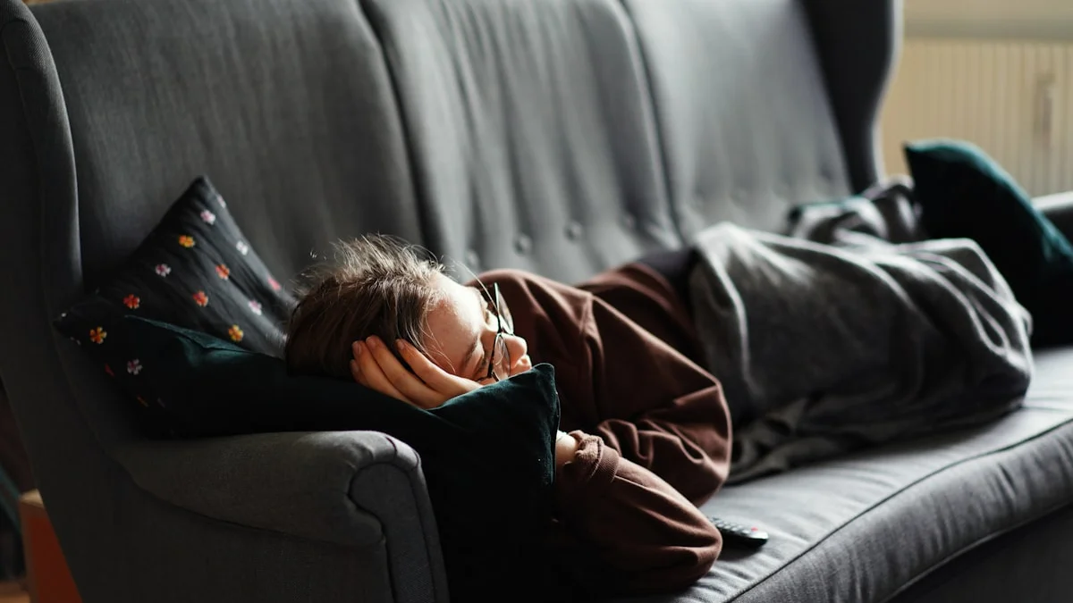 A woman lying flat on a grey couch under a blanket, hand resting over her face - a Quiet Core Reset guide to Are You Depleted or Just Tired? The 10-Minute Honest Energy Check.