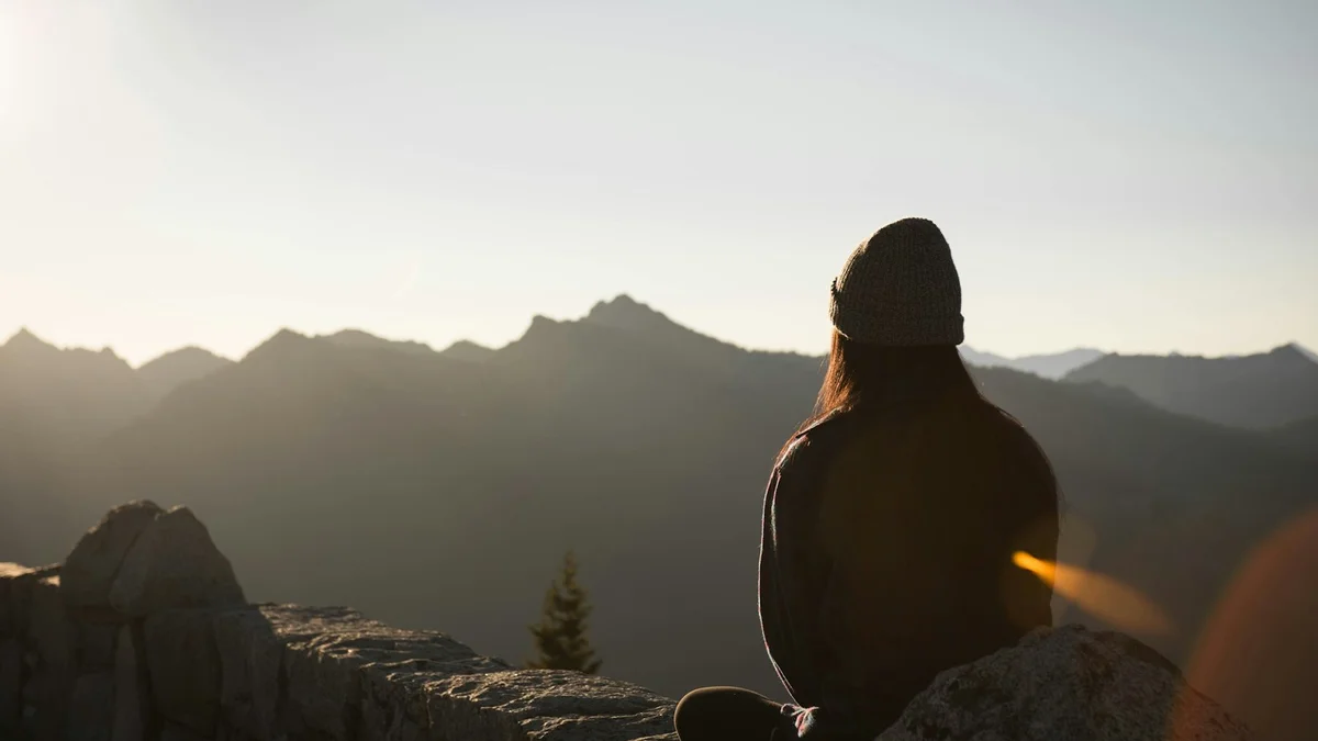 A person in a beanie sitting on a mountain edge, looking out over a sunlit range of distant peaks - a Quiet Core Reset guide to Digital Overload When You're Already Burned Out.