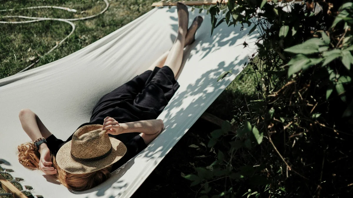 A person lying in a white hammock under leafy trees with a straw hat over their face - a Quiet Core Reset guide to Myth of Earned Rest.
