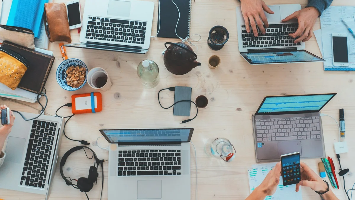 A top-down view of a busy shared table with laptops, phones, papers, headphones and coffee - a Quiet Core Reset guide to Energy Management for High Performers.