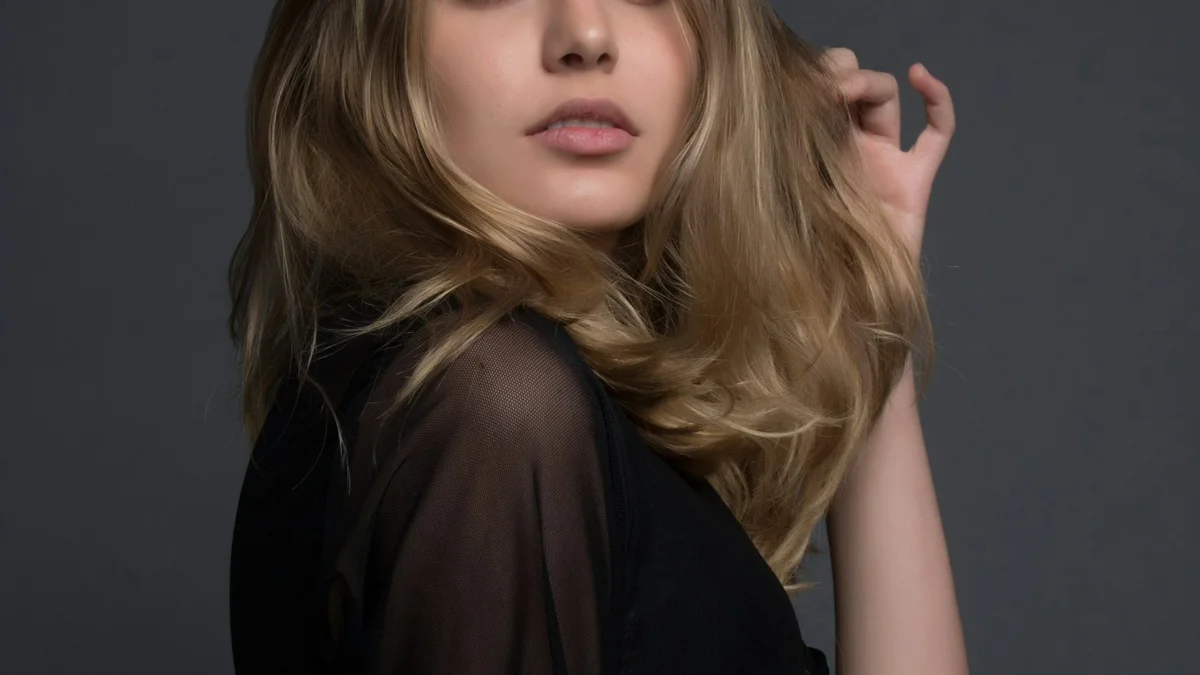 A portrait of a blonde woman in a dark sheer top, hand in her hair, on a grey background - a Quiet Core Reset guide to Are You an Energy Manager or a Time Manager? A 3-Minute Self-Check.