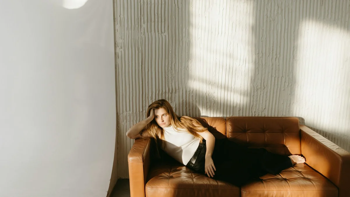 A tired woman resting on a leather couch in warm sunlight, letting her face do no work at all - a Quiet Core Reset guide to Forcing Positivity.