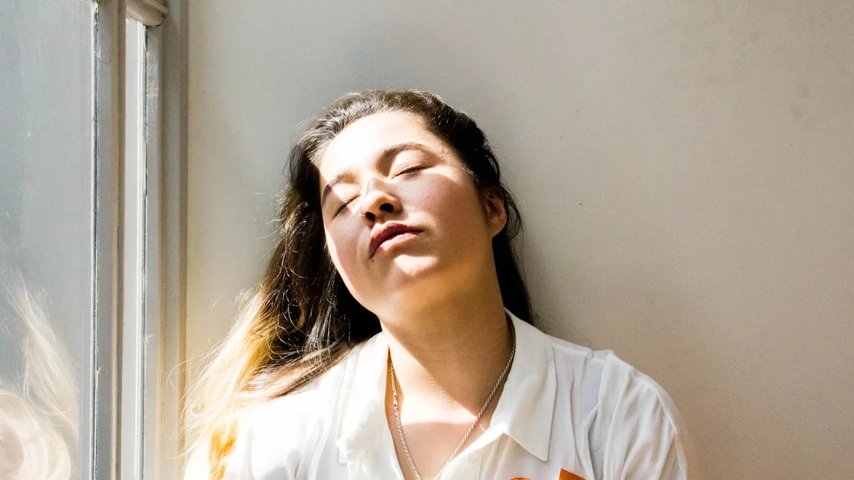 A woman resting with her eyes closed in warm sunlight beside a bright window - a Quiet Core Reset guide to How to Recharge Without Taking a Vacation.