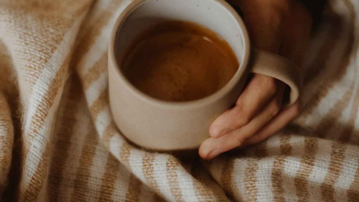 Two hands wrapped around a cream ceramic mug resting on a chunky striped blanket - a Quiet Core Reset guide to How to Say No Without Guilt.