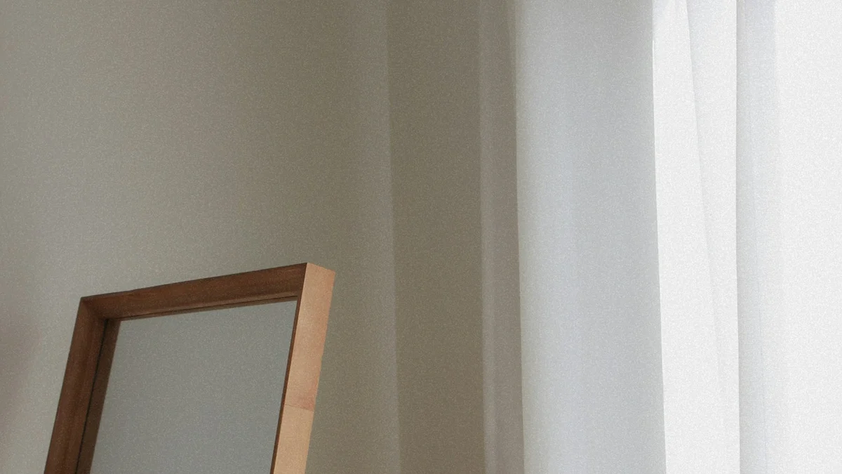 An empty wooden-framed mirror leaning beside a sheer curtain in soft daylight - a Quiet Core Reset guide to Inner Critic Explained Simply.