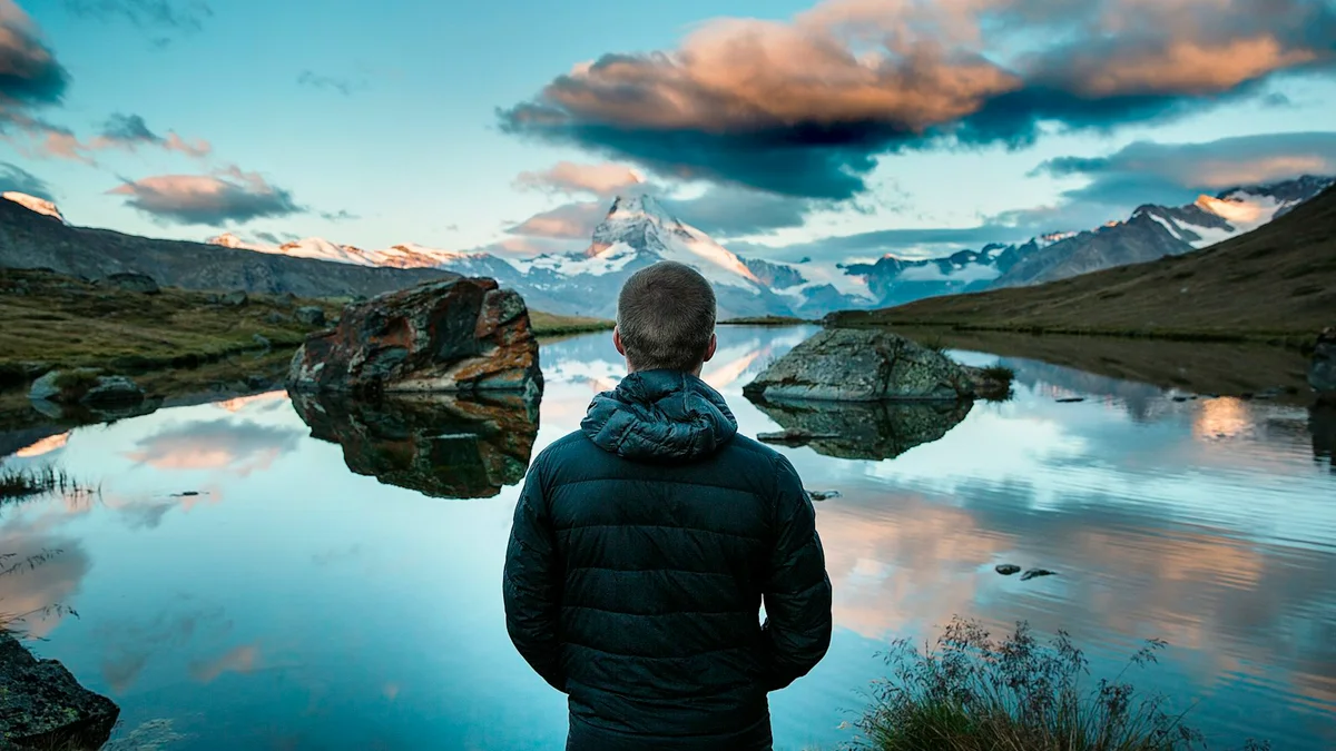 A person in a jacket standing before a still mountain lake reflecting snow-capped peaks - a Quiet Core Reset guide to Grayscale vs. Full Color.