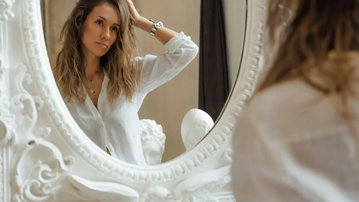 A woman rests a hand to her head as she studies her reflection in an ornate white mirror, soft neutral light - a Quiet Core Reset guide to Where Do You Feel It? - Locating Emotions in Your Body.