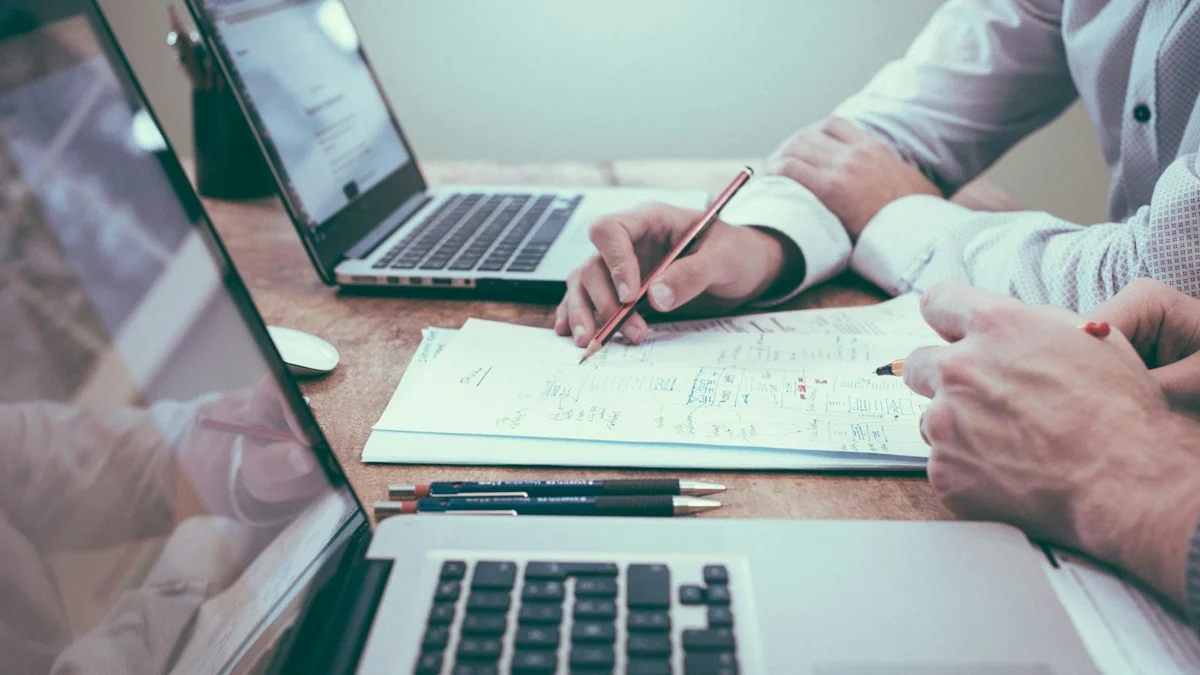 Two hands writing and pointing at a paper document on a desk with laptops, a planning meeting - a Quiet Core Reset guide to Micro-Reset Checklist.