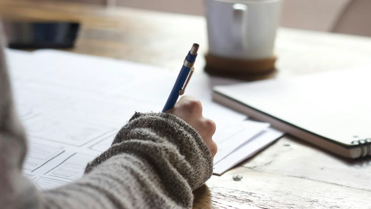 A hand in a knit sleeve writing with a blue pen on paper, a coffee cup nearby - a Quiet Core Reset guide to Are You Running Your Day Without Micro-Resets? A 3-Minute Self-Check.