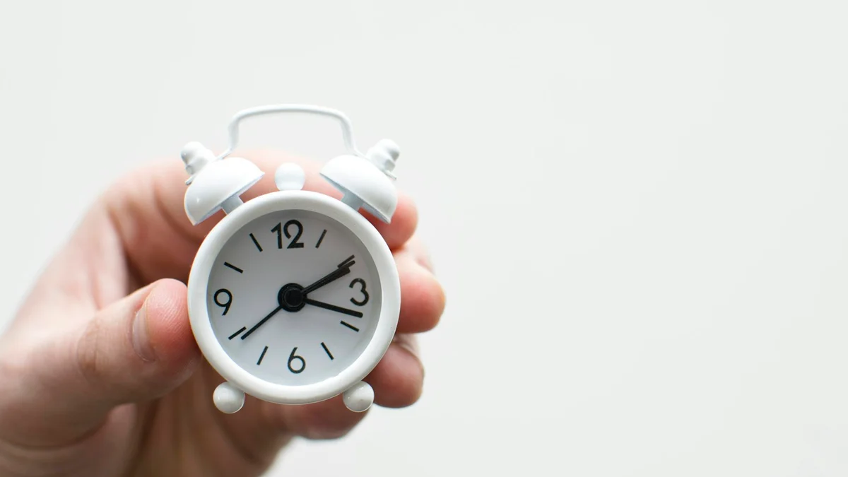 A hand holding a small white alarm clock against a plain background - a Quiet Core Reset guide to Morning Person vs. Night Owl.