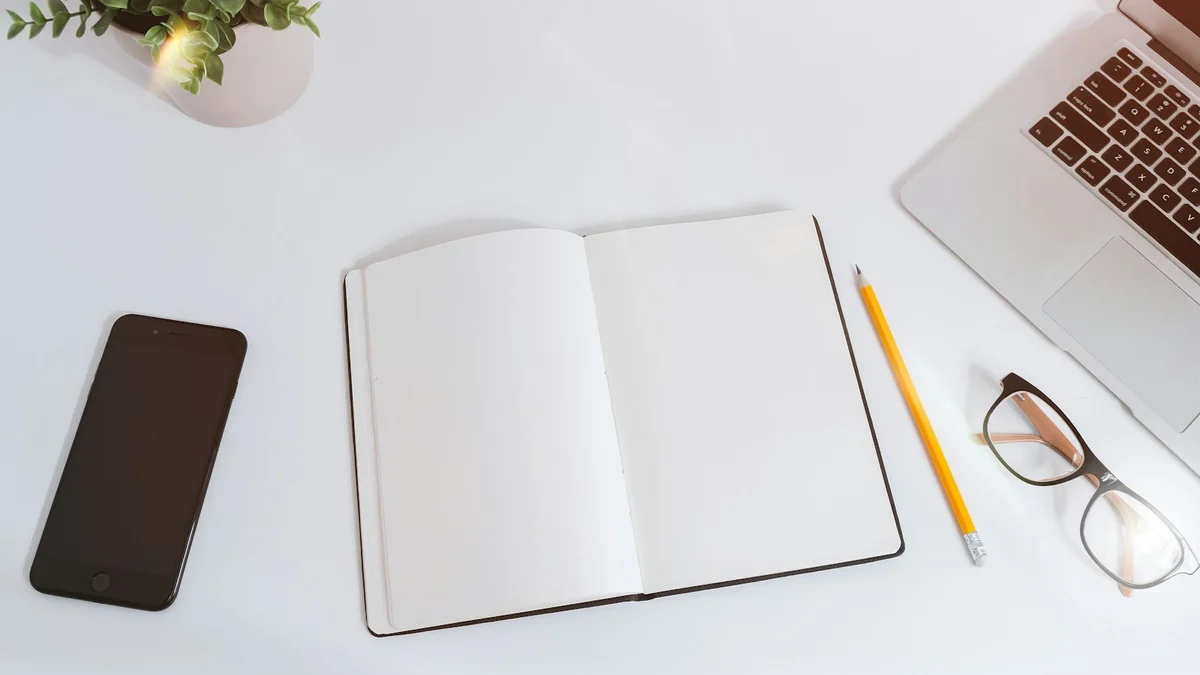 A clean white desk with an open blank notebook, a pencil, glasses, a phone and a laptop - a Quiet Core Reset guide to Peak Energy Planner.