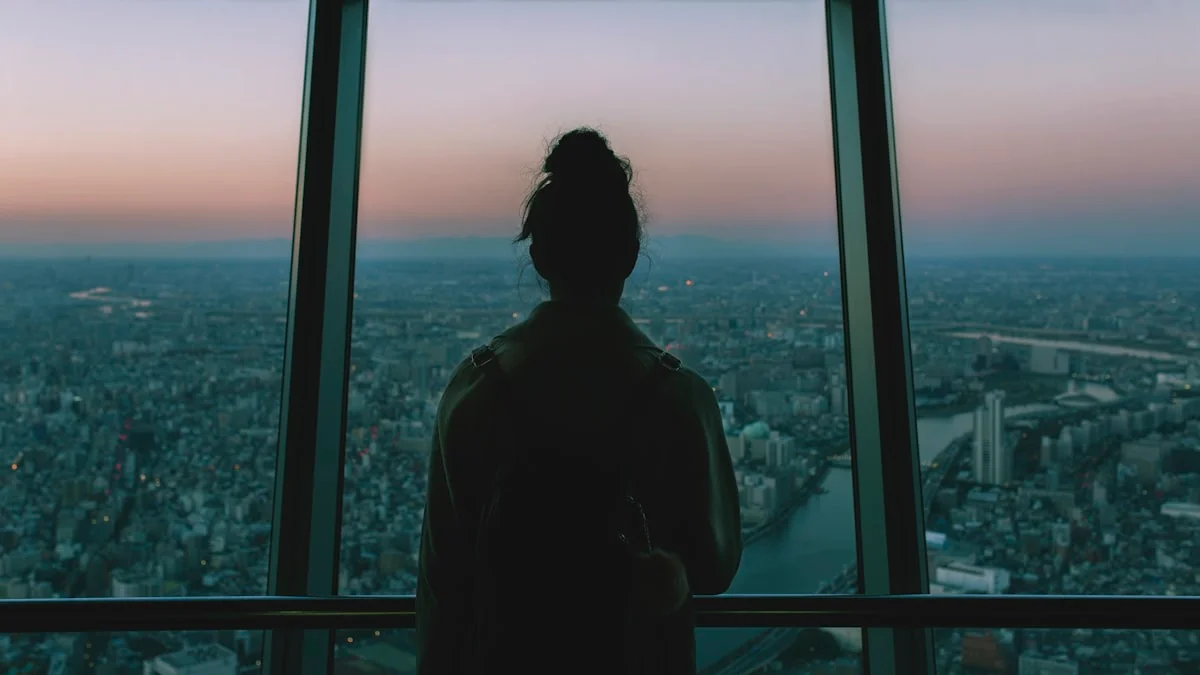 A woman seen from behind stands quietly at a vast window, watching the city fade into dusk - a Quiet Core Reset guide to Quiet Exhaustion vs. Burnout.