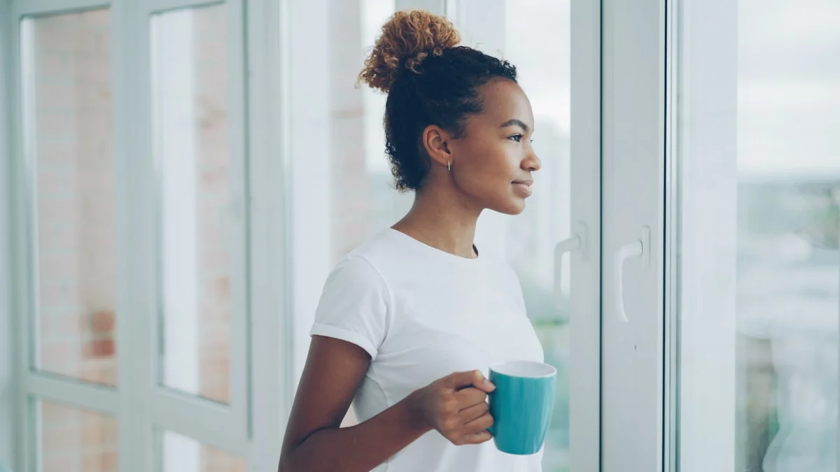 A woman in a white t-shirt holding a blue mug, pausing at a bright window to gaze outside - a Quiet Core Reset guide to Reclaiming Quiet Moments in a Noisy Feed.