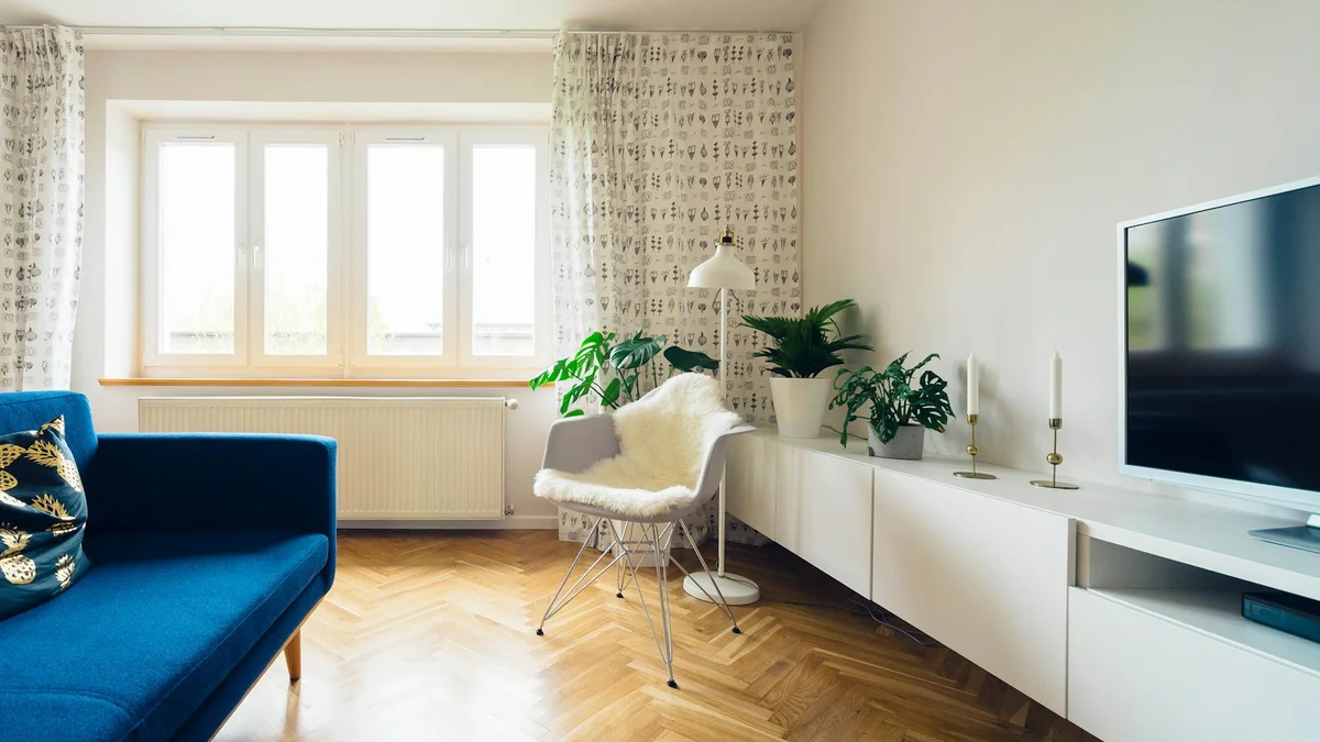 A cozy living room with a blue sofa, fresh plants, candles and a white fur chair by a bright window - a Quiet Core Reset guide to Why Rest and Digest Is Being Forgotten.