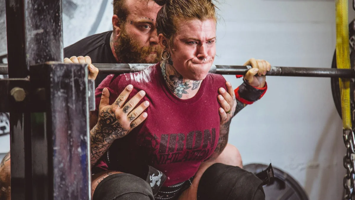 A woman straining under a heavy barbell squat in a gym, a man spotting her - a Quiet Core Reset guide to Rest & Digest for Low-Energy Days.
