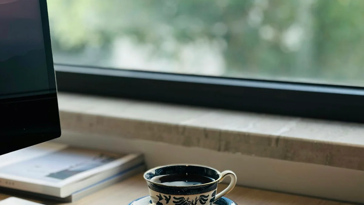 A cup of tea resting on an office desk beside a keyboard, with trees outside the window - a Quiet Core Reset guide to Rest Guilt at Work.