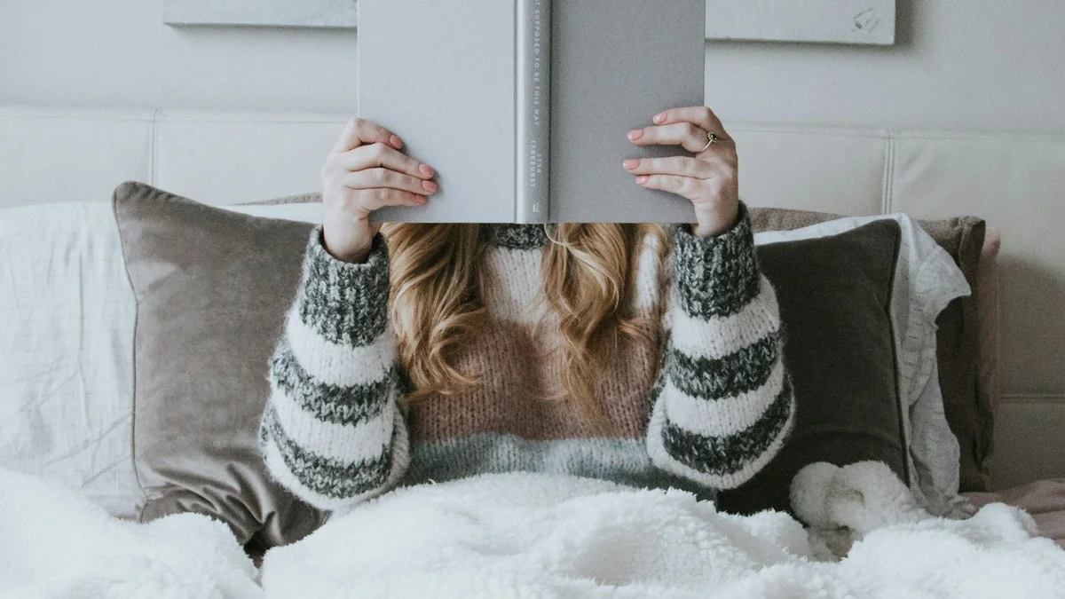A person reading in bed under a fluffy white blanket, holding the book above their face - a Quiet Core Reset guide to Beginner's Guide to Rest Guilt (and Unlearning It).