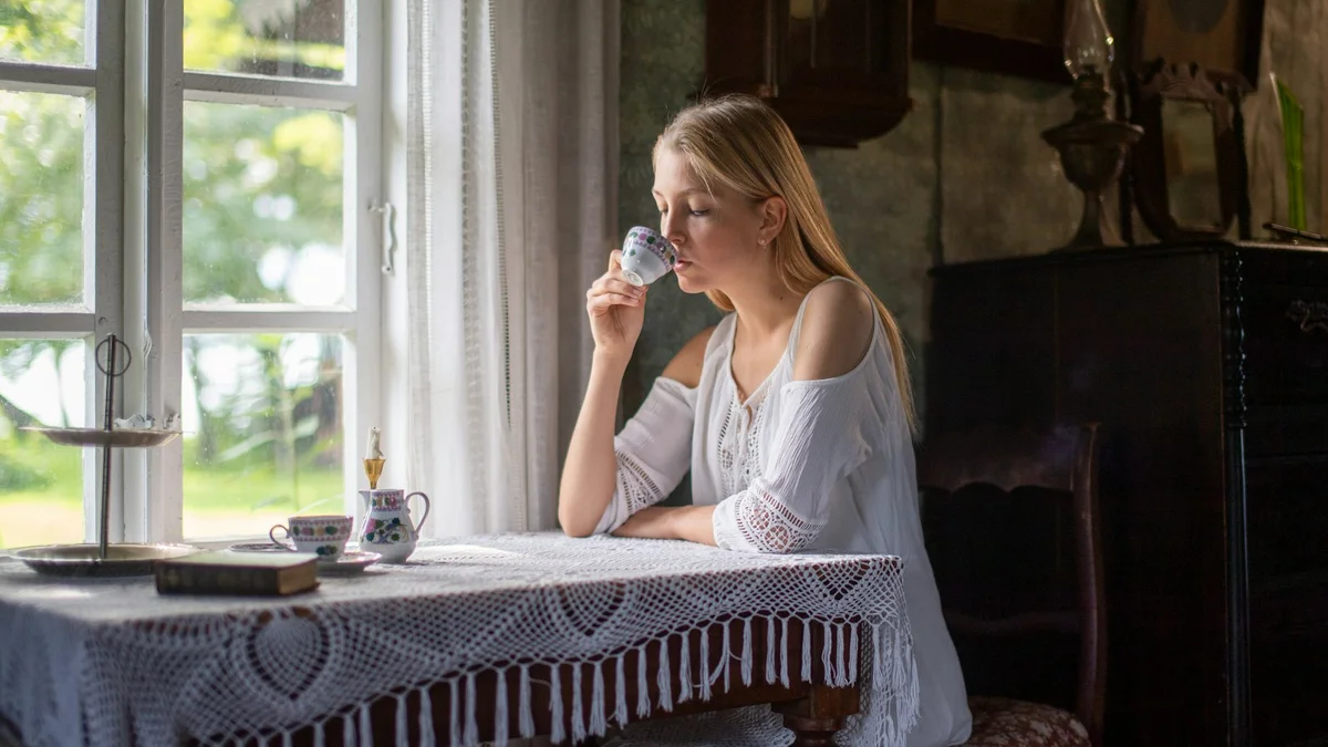 A woman in a white blouse sipping tea by a sunlit farmhouse window - a Quiet Core Reset guide to Rest Guilt for Moms.