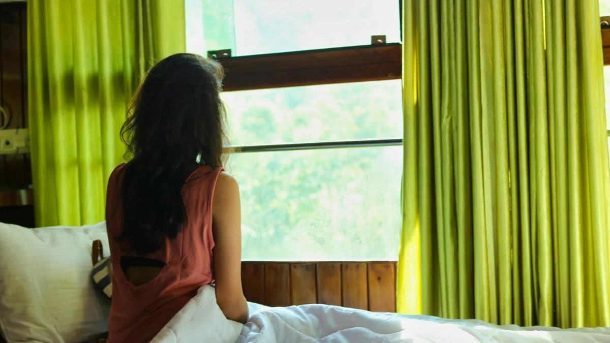 A woman sitting up in bed looking out a bright window - a Quiet Core Reset guide to 9 Signs Rest Guilt Is Running Your Life.