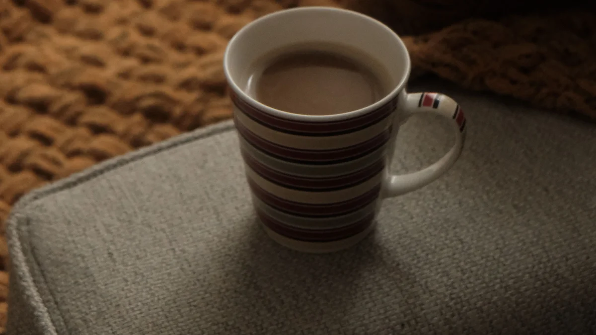 A striped mug of tea beside a chunky knit blanket on a couch arm in warm low light - a Quiet Core Reset guide to Sleep Recovery on Low-Energy Days.