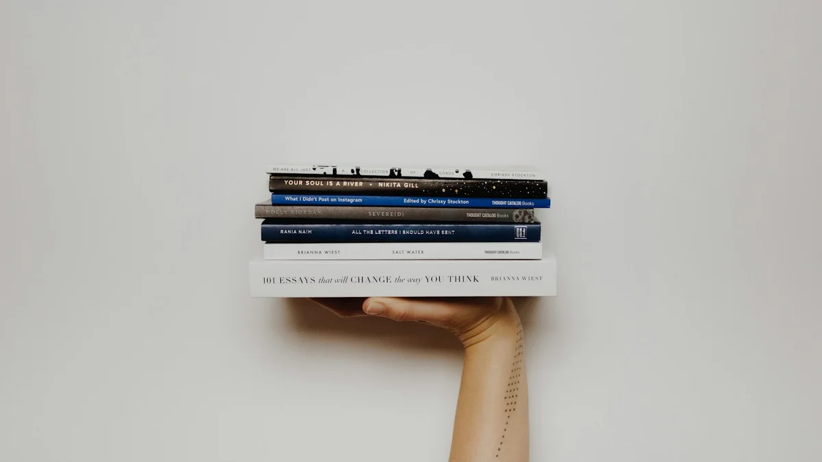A hand holding a stack of books against a plain wall, one arm raised - a Quiet Core Reset guide to Slow Living for High Performers.