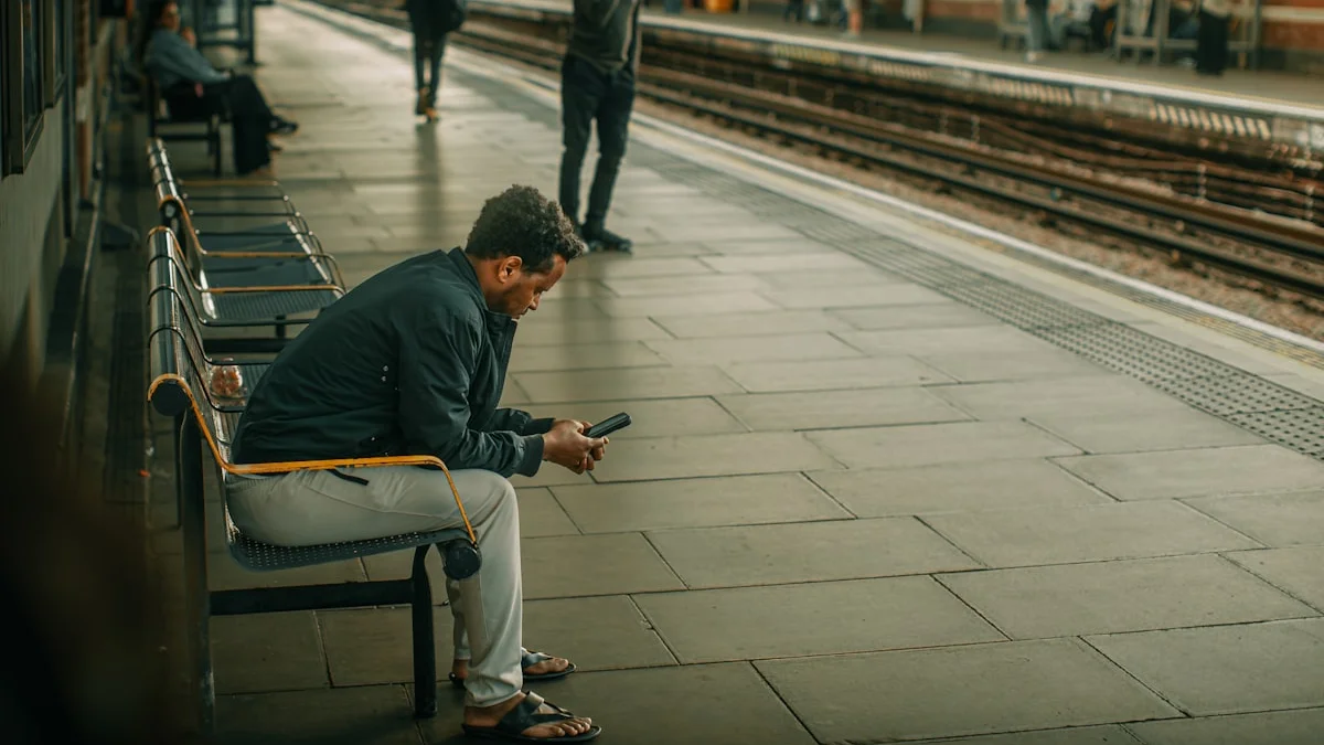 A man hunched alone on a train-platform bench, scrolling his phone while he waits - a Quiet Core Reset guide to Subtle Signs Hustle Culture Is Draining You Quietly.