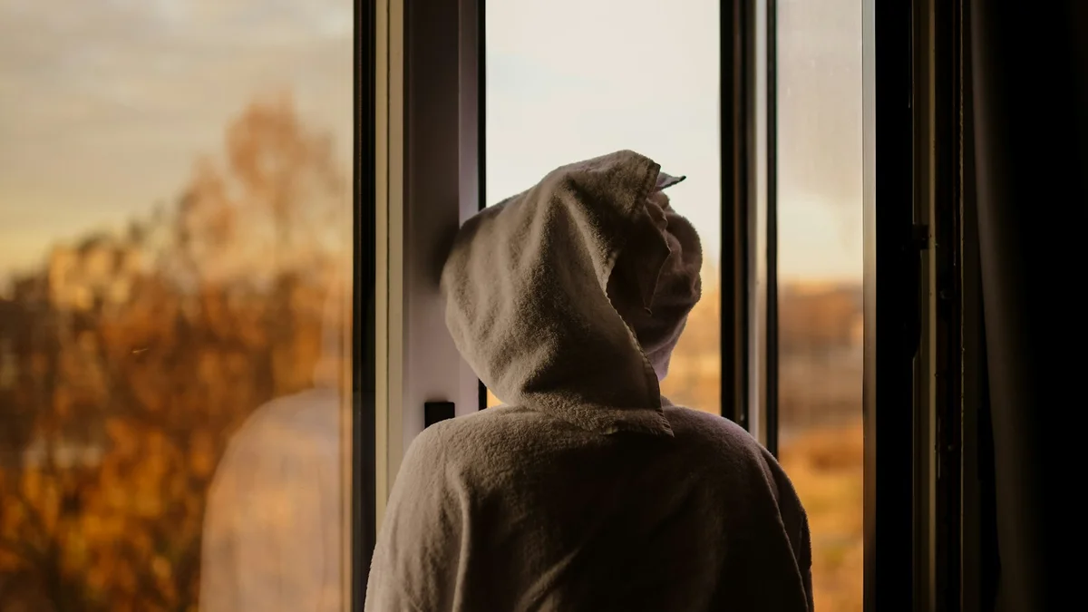 A person in a hooded robe looking out a window at golden trees - a Quiet Core Reset guide to Survival Mode at Home.