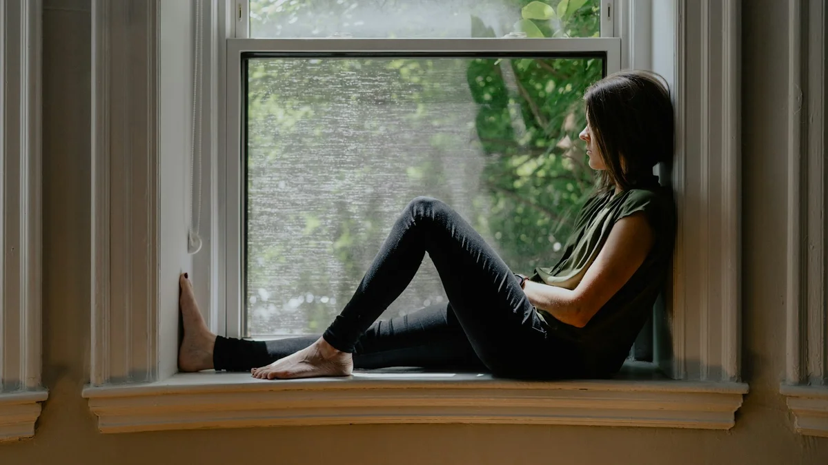A woman seated on a wide window sill gazing out at green trees in soft morning light - a Quiet Core Reset guide to What Is the Inner Critic? The Voice Behind Your Exhaustion.