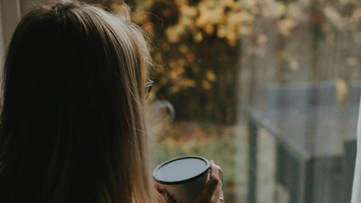 A woman holding a warm cup of coffee, gazing out a bright window with no phone in hand - a Quiet Core Reset guide to why boredom is good for your nervous system.