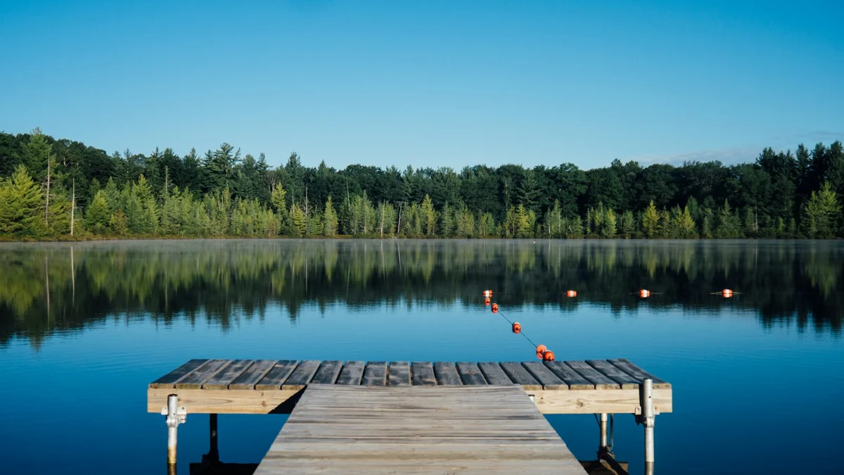 A wooden dock stretching over a calm blue lake with forest on the far shore and a clear sky - a Quiet Core Reset guide to Why Resting Proactively Prevents Chronic Exhaustion.