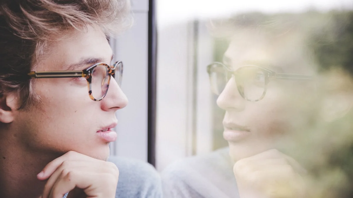A man resting his chin on his hand, gazing pensively through a window with his reflection - a Quiet Core Reset guide to why you can't just relax.