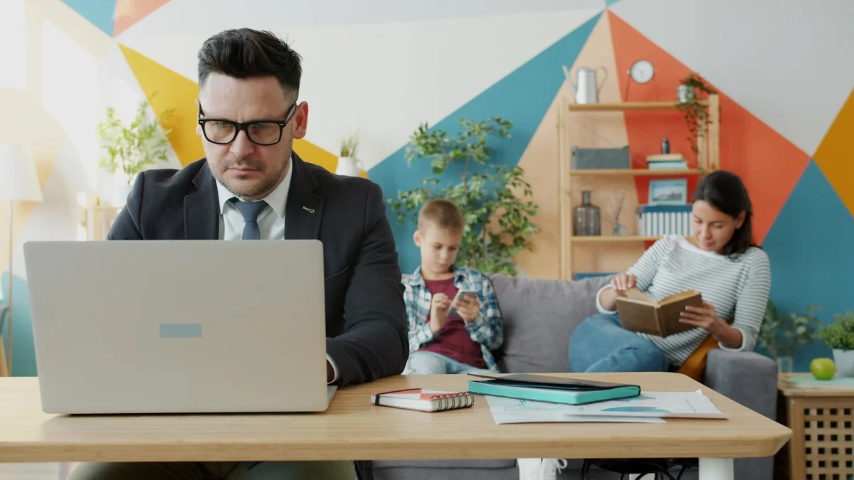 A father working on a laptop at home while his family relaxes behind him - a Quiet Core Reset guide to Work Energy for Parents.